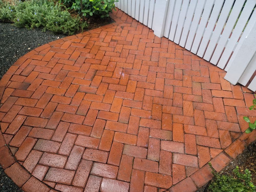 Wet brick pathway with a herringbone pattern leading to a white picket fence.