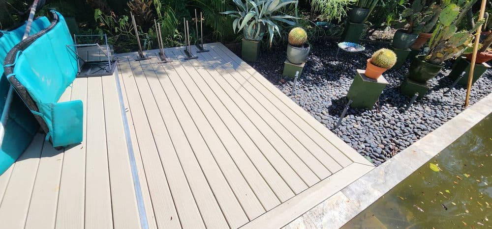 Sunlit deck with seating, surrounded by potted plants and decorative stones near water.