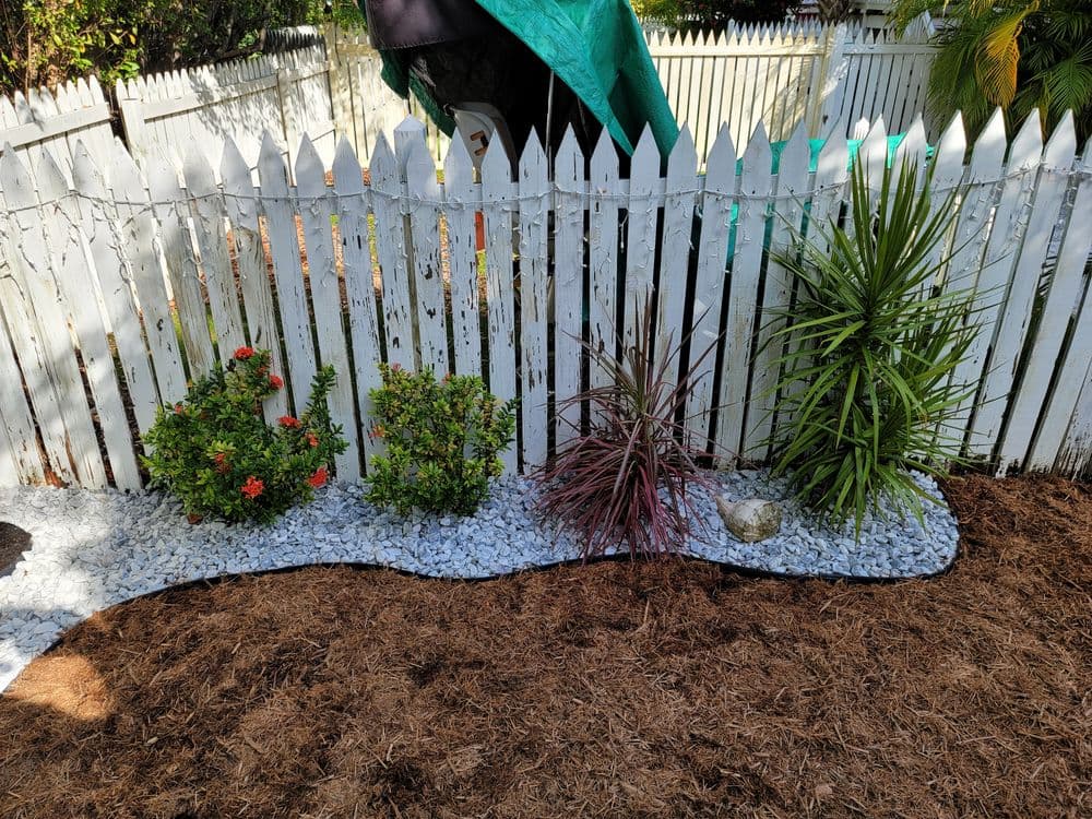 Lush garden with colorful plants and white gravel along a white picket fence.