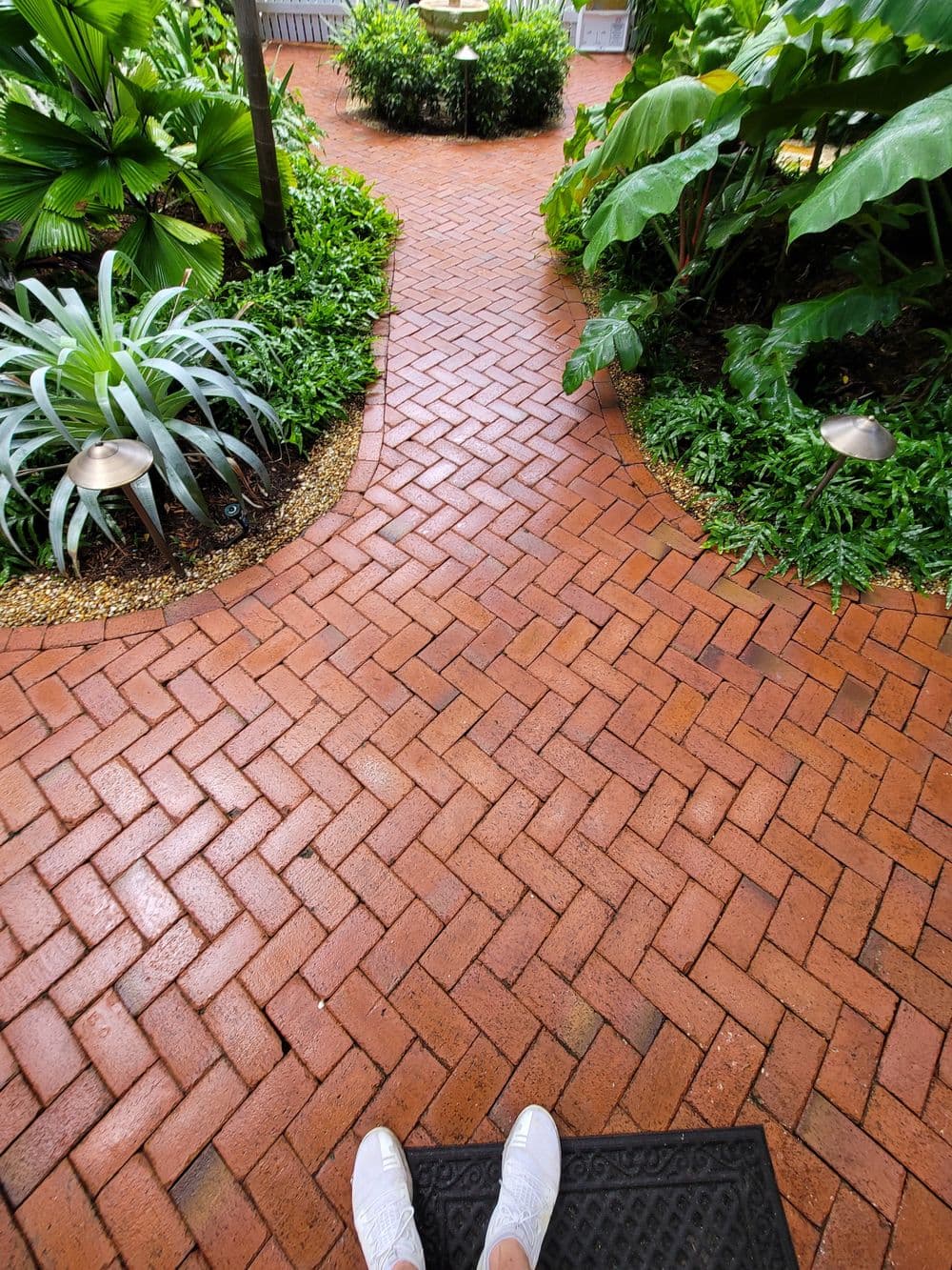 Pathway made of red brick surrounded by lush greenery and decorative lighting.