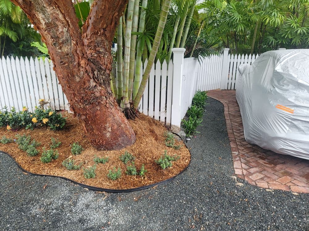Tree beside a white picket fence with landscaped plants and a covered car on a brick path.