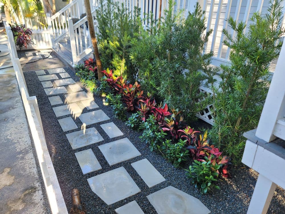 Lush garden pathway with stone steps, vibrant plants, and greenery beside a residential walkway.
