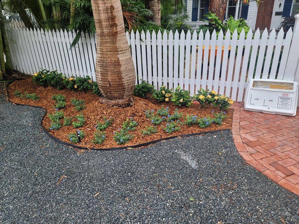 Flower bed with blooming yellow and blue flowers surrounded by mulch and a white picket fence.