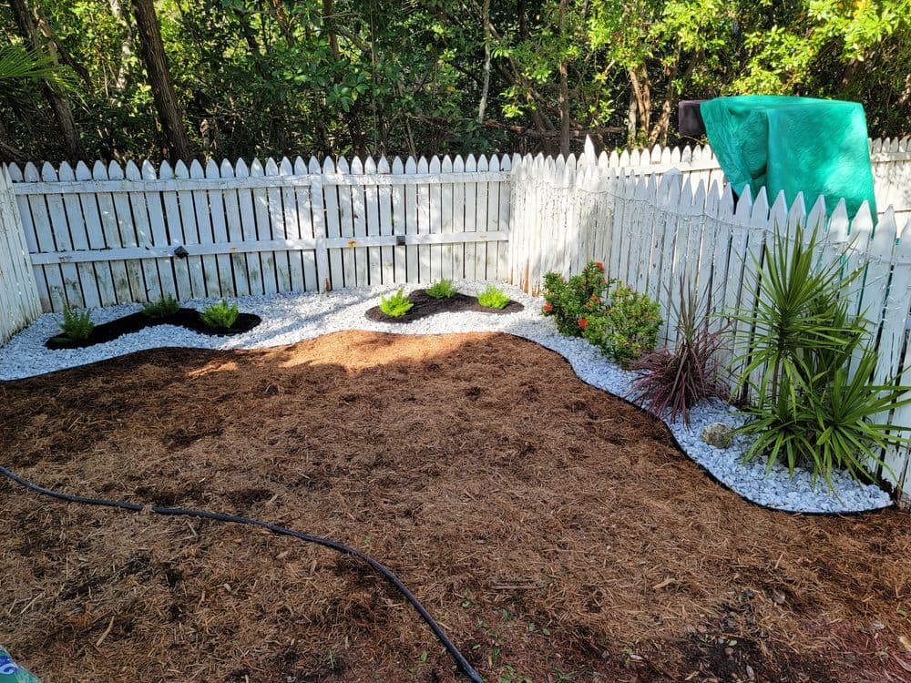 Landscaped garden area with white gravel, plants, and a white picket fence in a sunny setting.